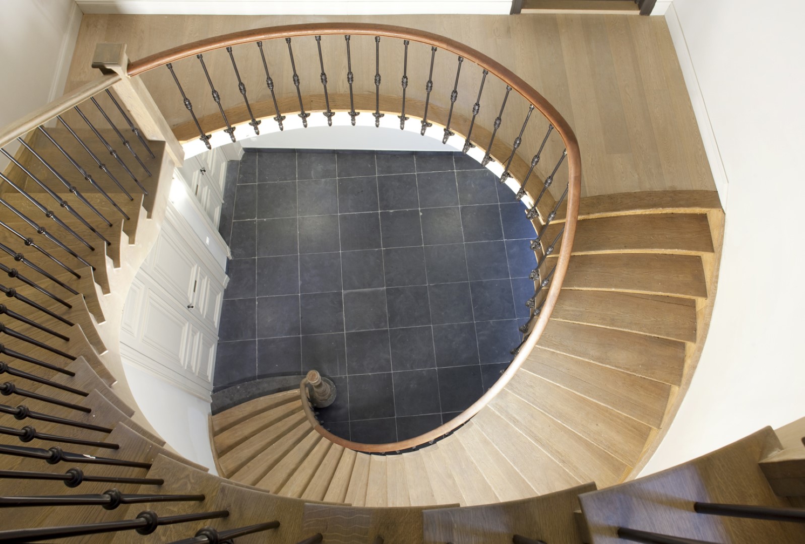 Elegante houten trap in een spiraalvorm met zwarte smeedijzeren spijlen en houten leuning, uitkomend op een vloer met grote donkere tegels.