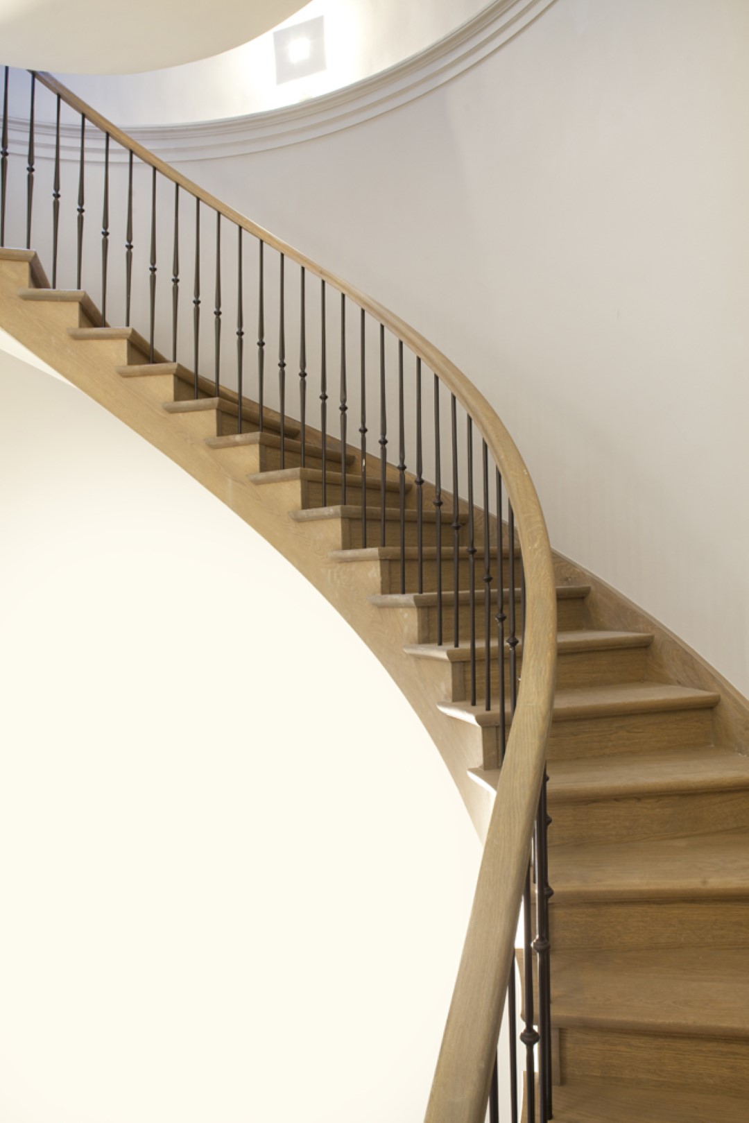 Houten trap met sierlijke bocht, zwarte spijlen en houten leuning, oplopend langs een gladde witte muur in een lichte ruimte.