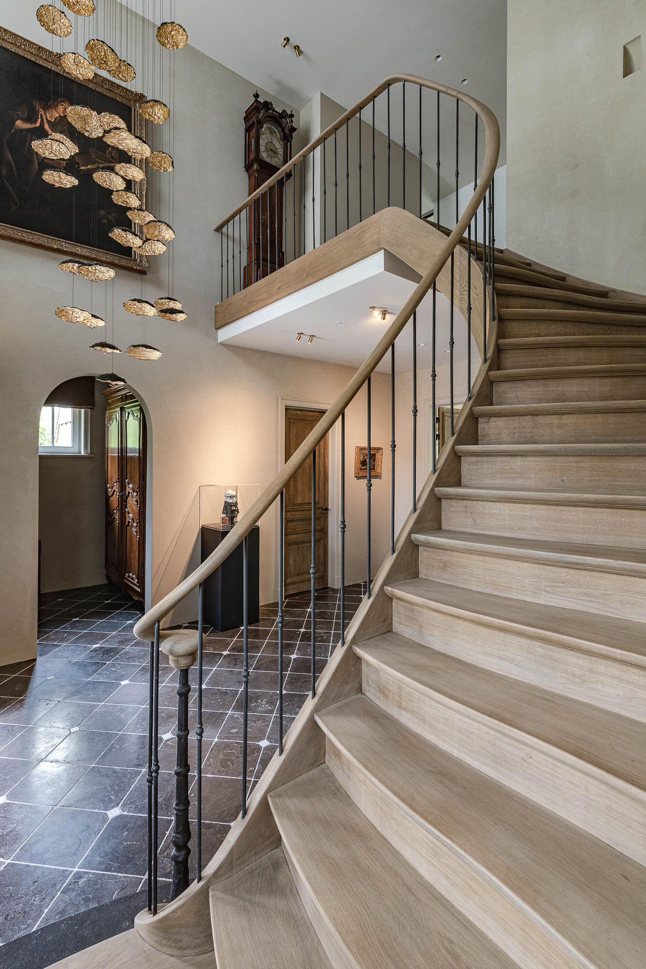 Een imposante, licht gebogen houten trap met zwarte spijlenleuning voert omhoog in een ruime hal met donkere tegelvloer, hoge plafonds en een opvallende hanglampinstallatie; klassieke elementen zoals een staande klok en boogdeur contrasteren met het moderne interieur.