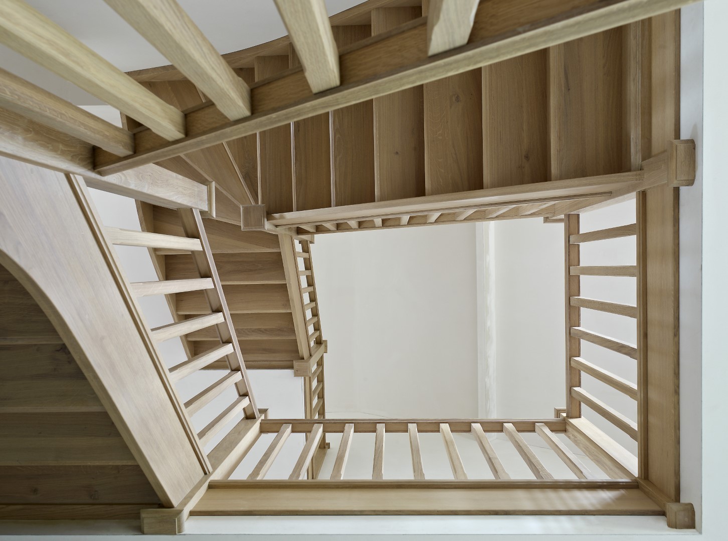 Uitzicht naar boven door het trapgat van een houten trap met rechte spijlen en gesloten treden, in een lichte ruimte met witte muren.