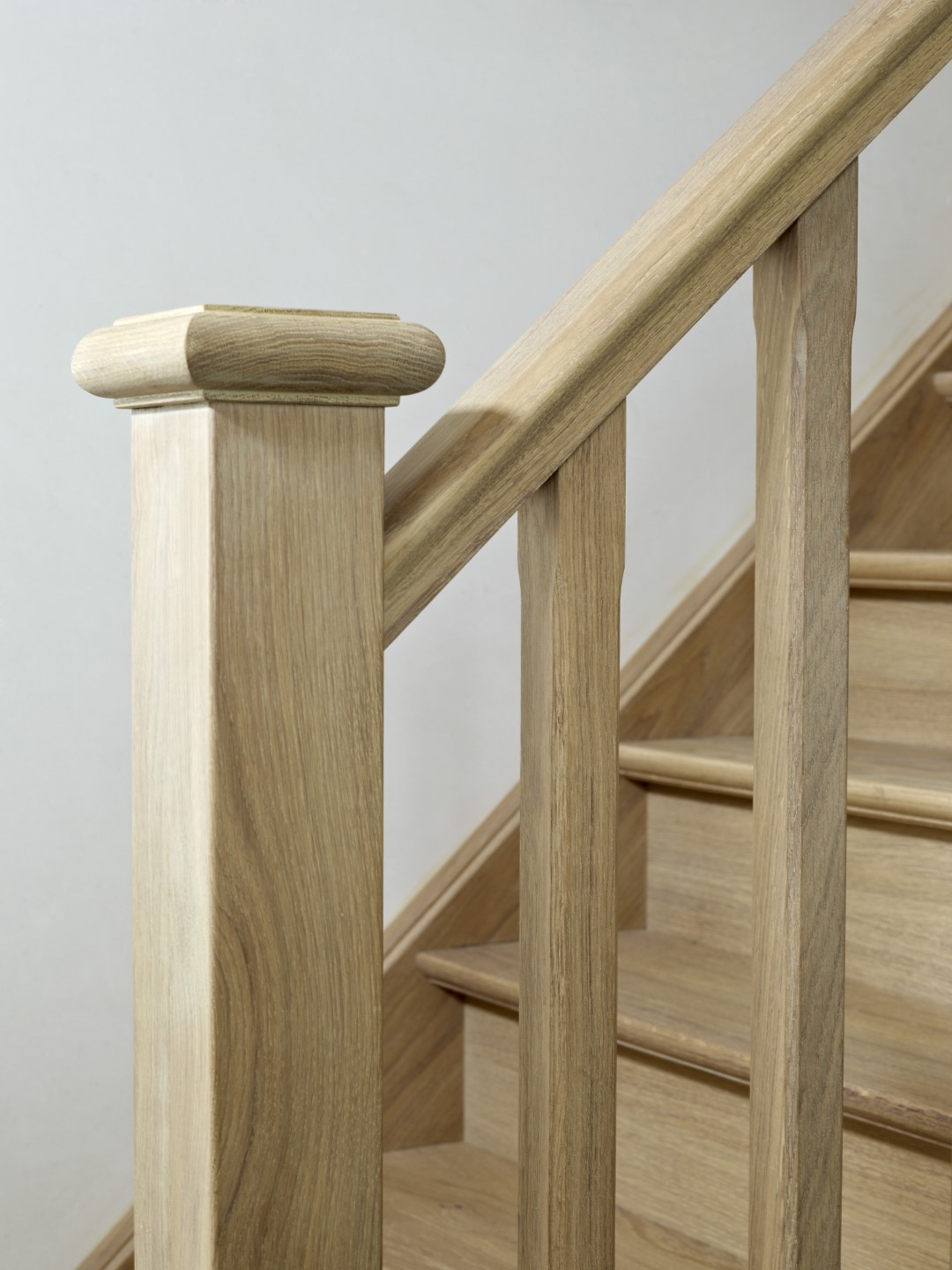 Close-up van een massieve houten trap met rechte spijlen, gladde leuning en afgeronde baluster in een lichte ruimte.