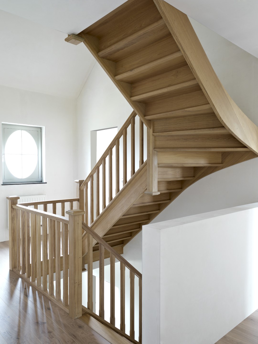 Trap in massief hout met rechte balustrade en gesloten treden, geplaatst in een lichte ruimte met witte muren, een rond raam en houten vloer.