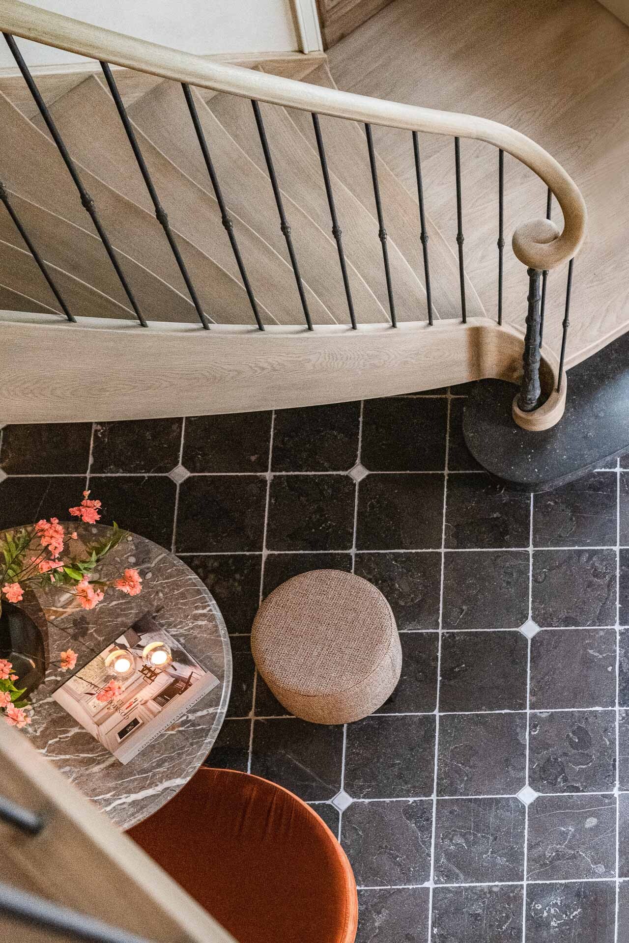 Een sfeervol bovenaanzicht van een hal met een sierlijk gebogen houten trapleuning en zwarte spijlen, een zwart marmeren tegelvloer, een ronde marmeren tafel met bloemen en tijdschriften, en twee ronde zitpoefs in verschillende stoffen.