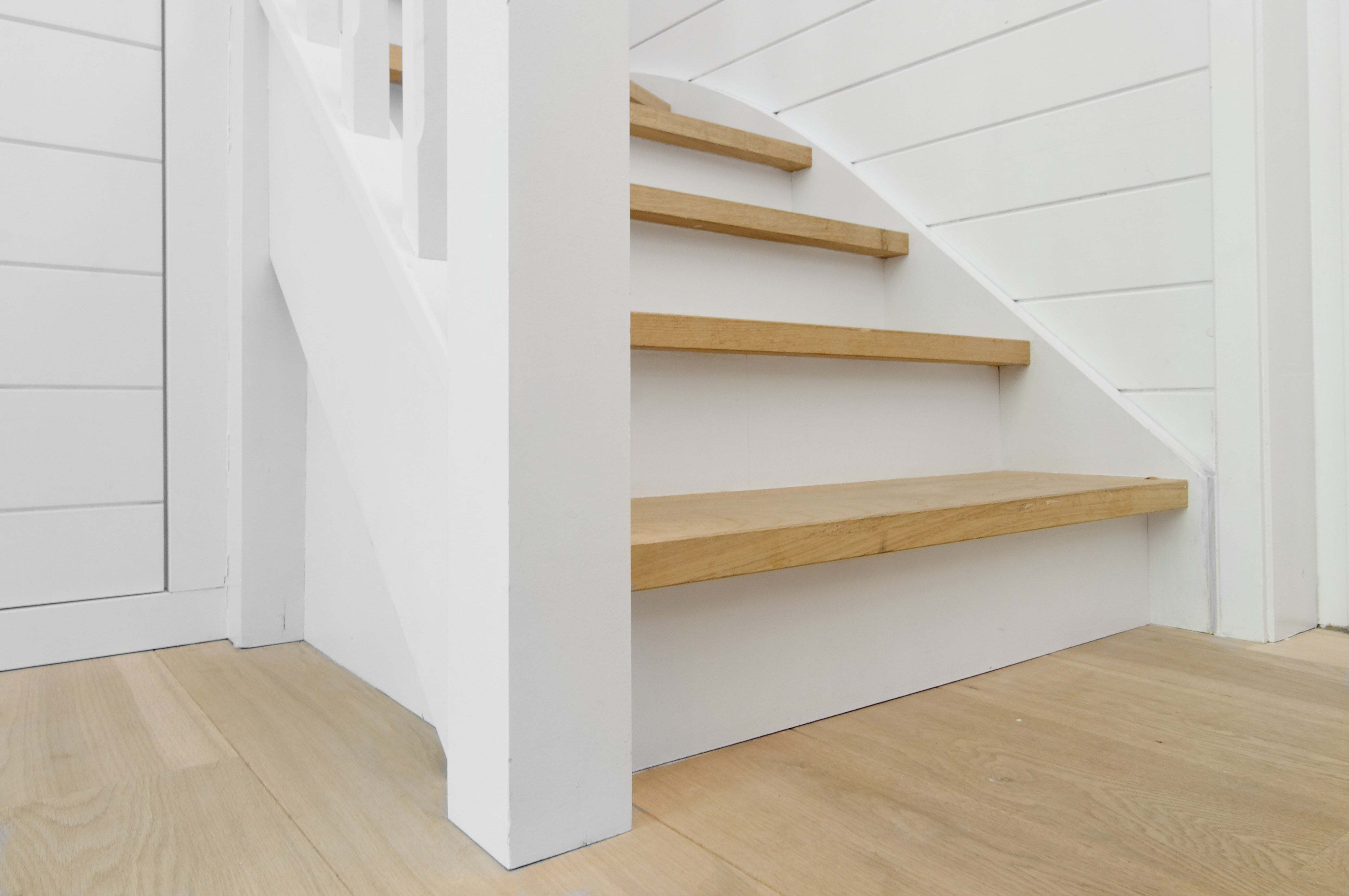 Een close-up van de onderzijde van een witte houten trap met naturel houten treden, geplaatst in een licht interieur met wit lambrisering op de muur en een licht houten vloer.