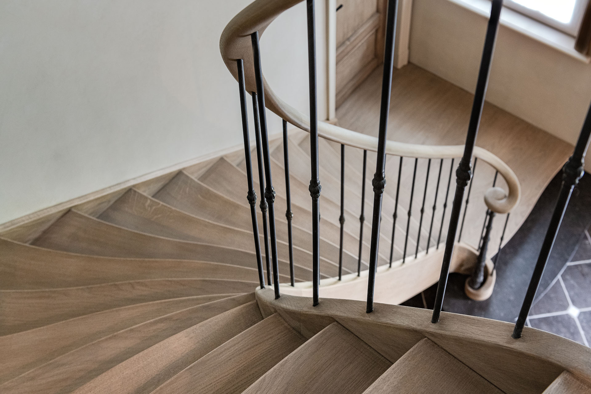 Een bovenaanzicht van een elegant gebogen houten trap met sierlijke zwarte spijlen en een afgeronde houten leuning, die afdaalt naar een hal met een donker betegelde vloer en een groot raam aan de zijkant.