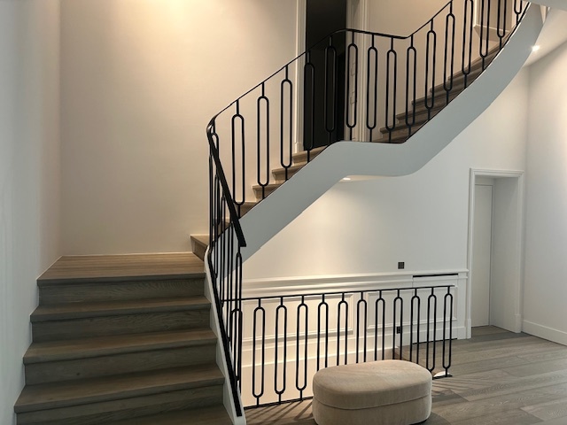 Een stijlvolle trap met lichtgrijze treden, een witte zijkant en een zwart metalen leuning met geometrisch patroon, naast een ronde beige poef.