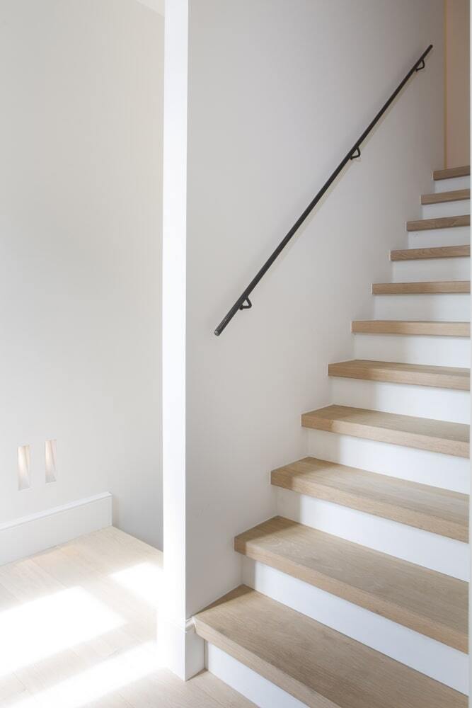 Een moderne trap met lichte houten treden en witte stootborden, geplaatst in een helder en minimalistisch interieur; aan de muur rechts loopt een slanke zwarte metalen leuning omhoog, met invallend daglicht op de vloer.