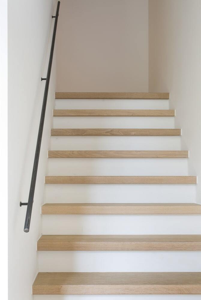 Een eenvoudige, strak vormgegeven trap met lichte houten treden en witte stootborden, tegen een neutrale muur; aan de linkerzijde loopt een slanke zwarte metalen leuning omhoog.