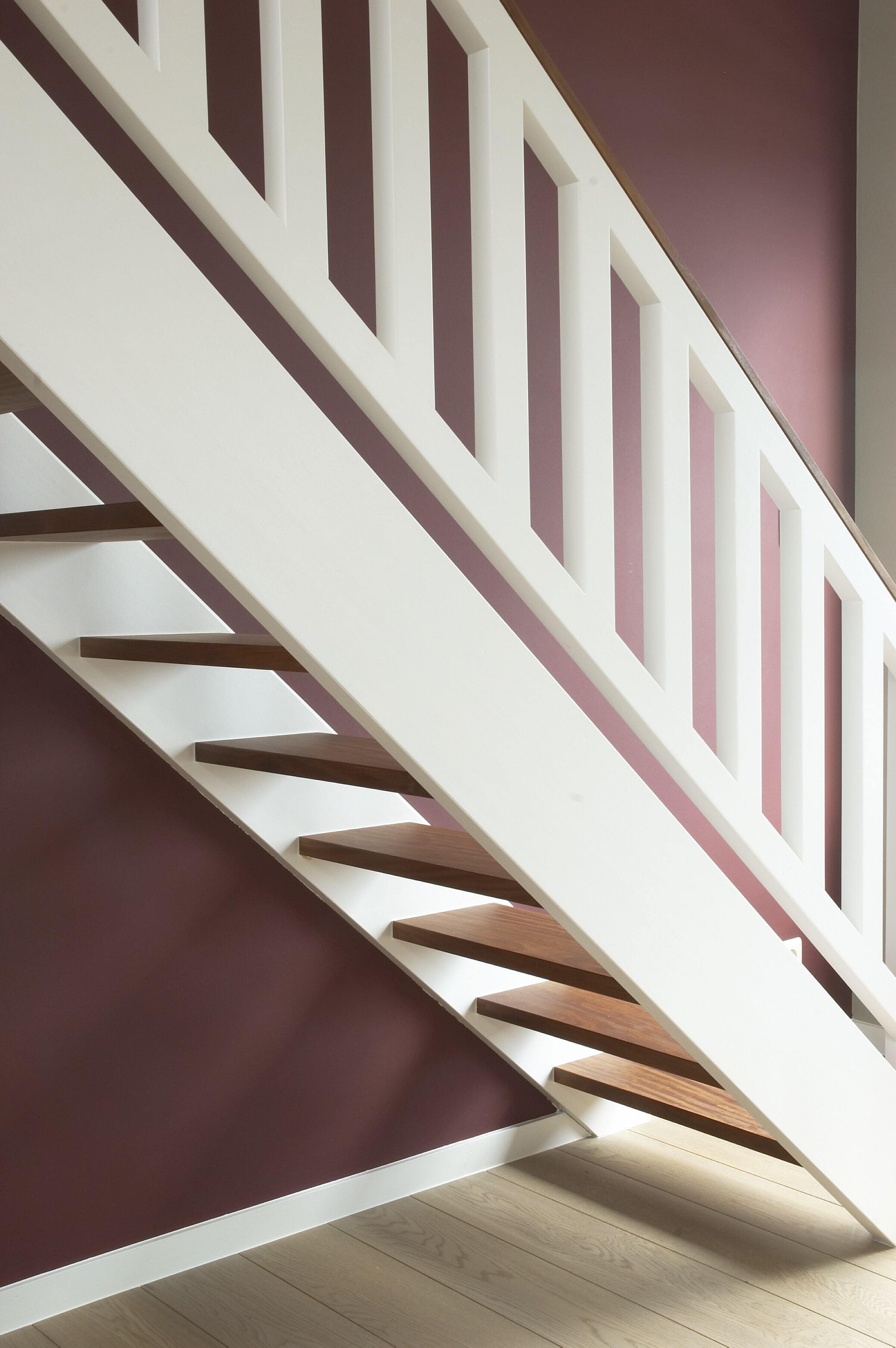 Een moderne open trap met witte zijkanten en leuning, voorzien van contrasterende donkere houten treden; de trap staat tegen een auberginekleurige muur en rust op een licht houten vloer.