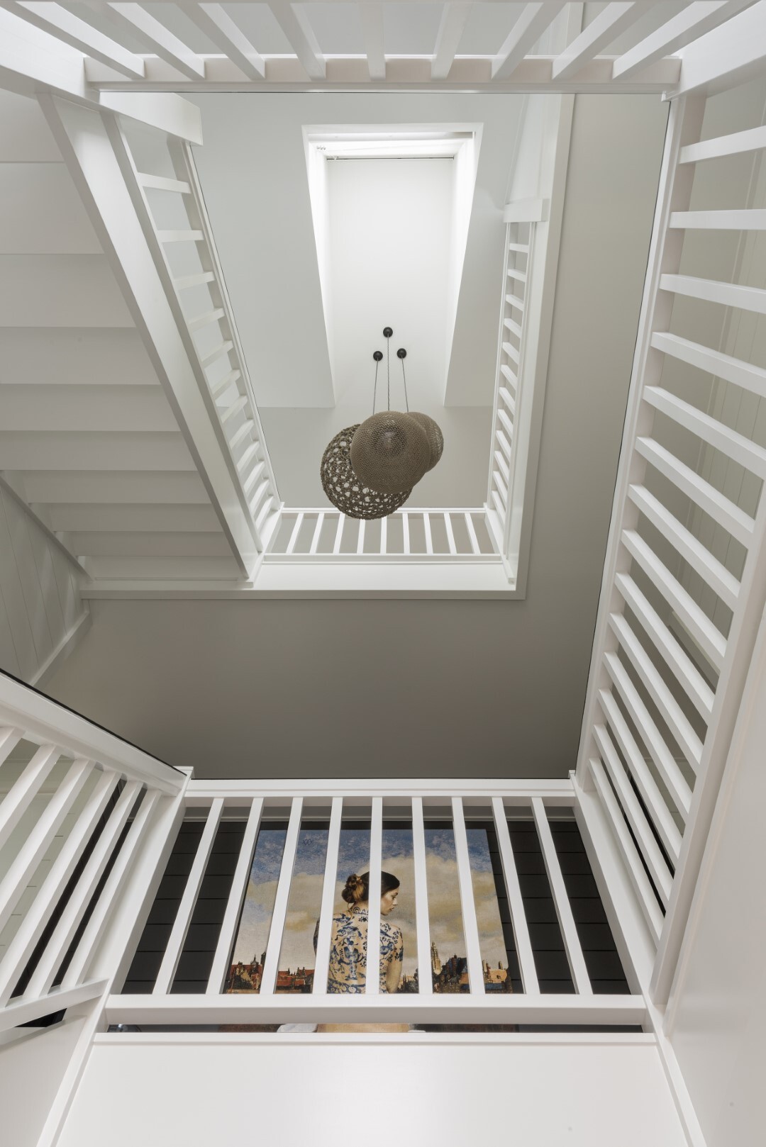 Doorkijk naar boven in een witte trappenhal met meerdere verdiepingen, een ronde hanglamp in het midden en een schilderij zichtbaar op de onderste etage.