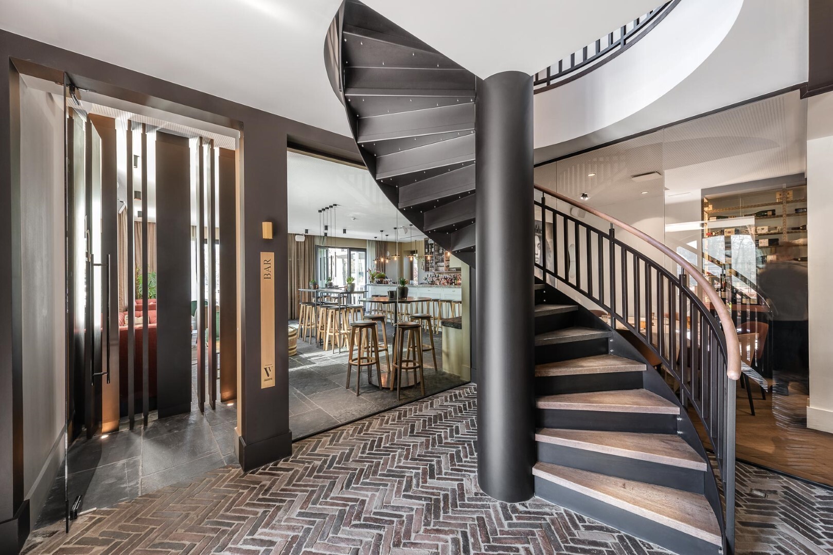 Een zwarte spiraaltrap in een stijlvol, modern interieur met visgraatvloer en een open zicht op een eet- en zithoek.