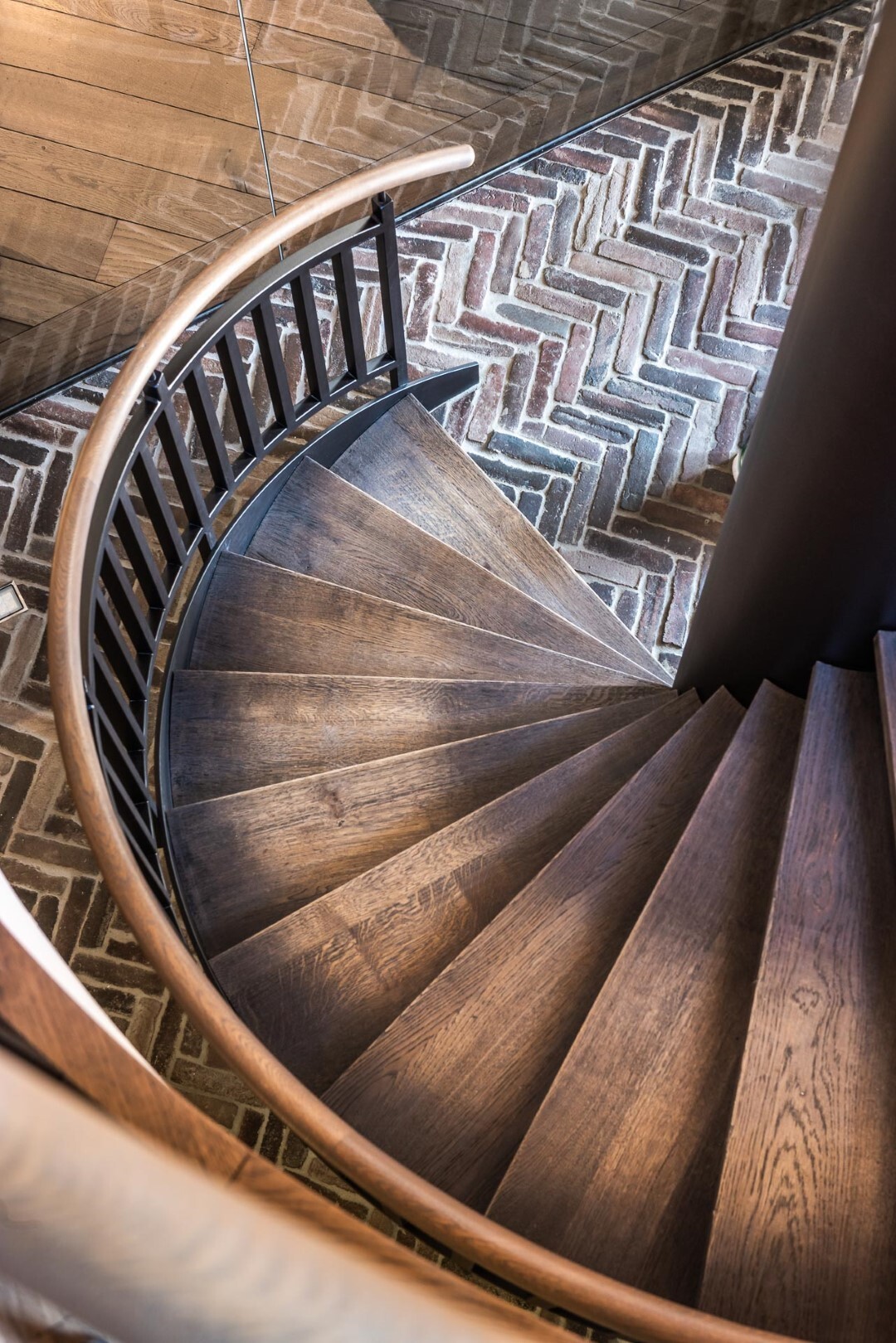 Een spiraaltrap met donkerhouten treden en een gebogen leuning, uitkomend op een visgraatvloer in aardetinten.
