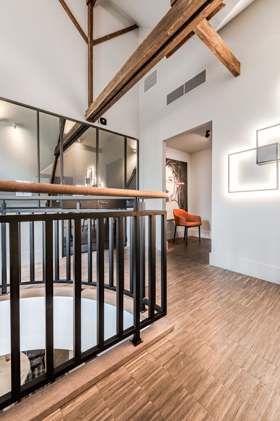 Een moderne overloop met zwarte balustrade rond een vide, houten plafondbalken, glazen wand, en zicht op een oranje stoel in de gang.