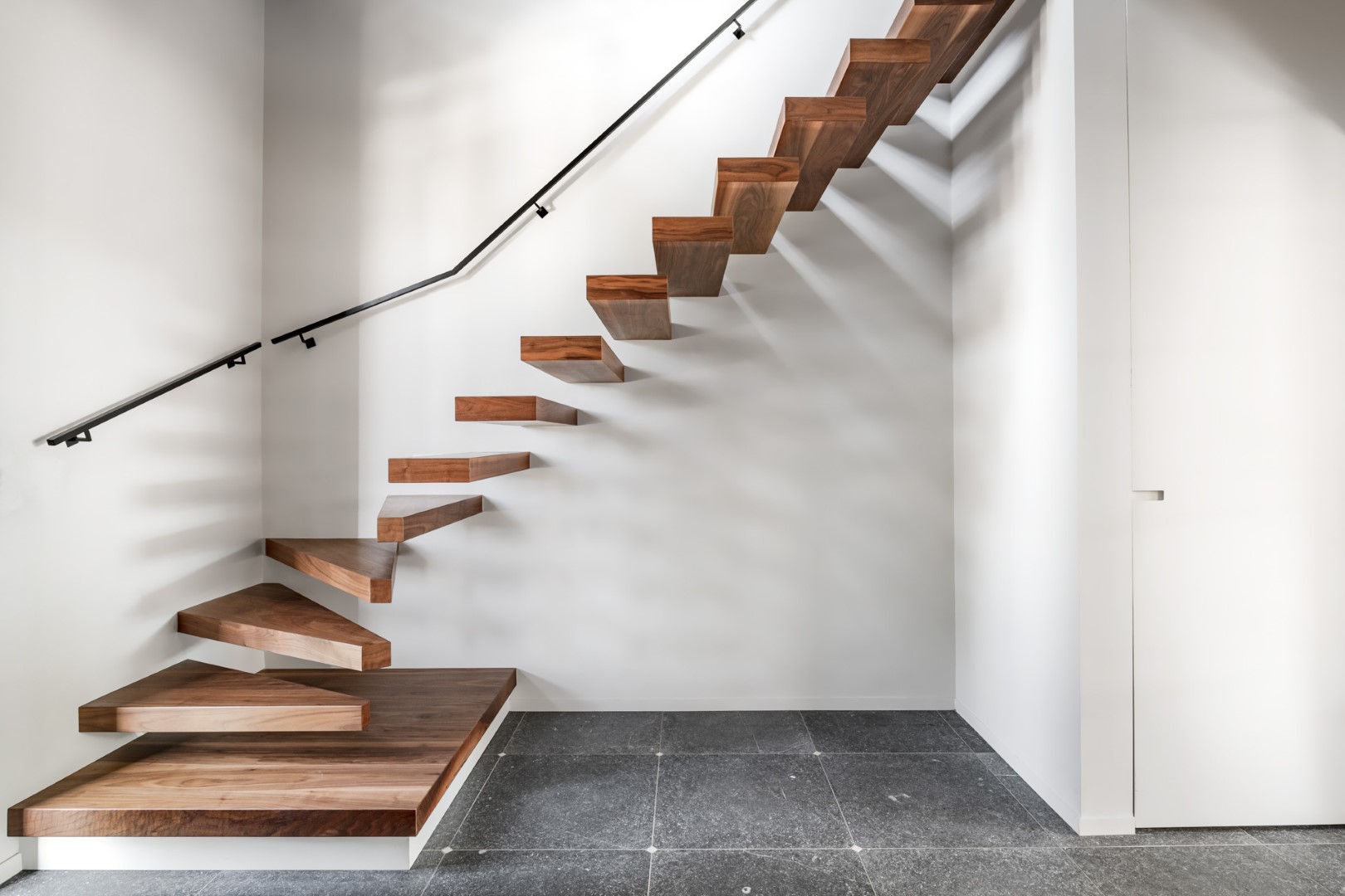 Moderne zwevende houten trap met donkere treden tegen een witte muur, voorzien van een zwarte metalen leuning en geplaatst boven een grijze tegelvloer.
