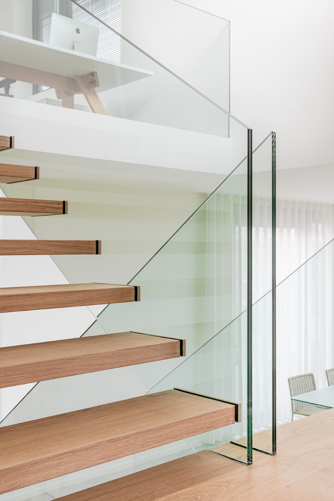 Trap met zwevende houten treden en strakke glazen balustrade, in een lichte moderne ruimte met witte gordijnen en een bureau op de bovenverdieping.
