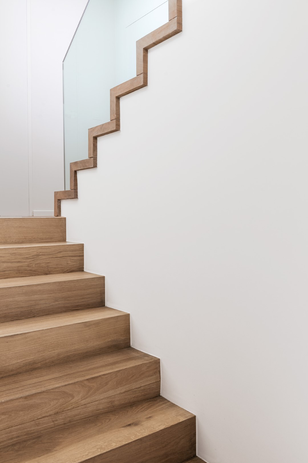 Moderne trap met brede houten treden en een glazen balustrade met houten rand die in een zigzaglijn het verloop van de trap volgt.