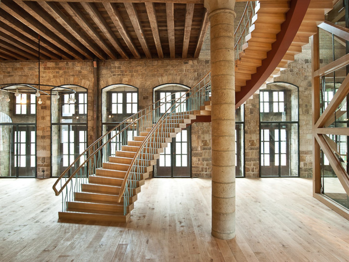 Grote houten trap met golvende metalen spijlen en houten leuning in een ruime hal met stenen muren, hoge ramen, houten balkenplafond en licht houten vloer.