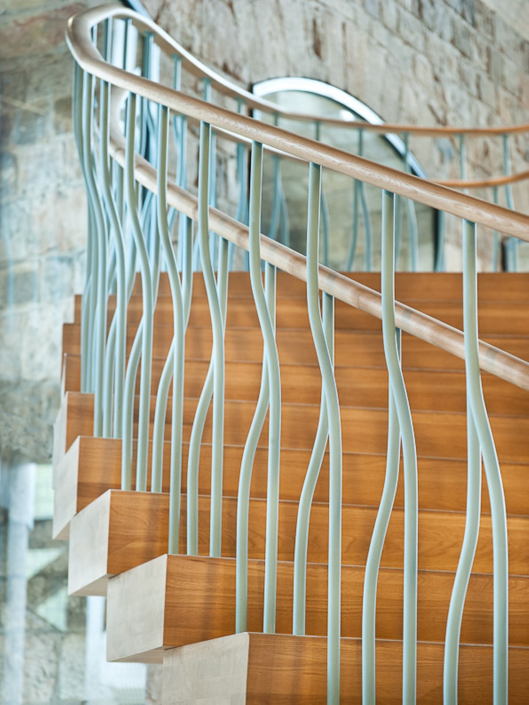 Trap met warme houten treden en lichtblauwe golvende metalen spijlen, afgewerkt met een gladde houten leuning, tegen een muur van natuursteen.