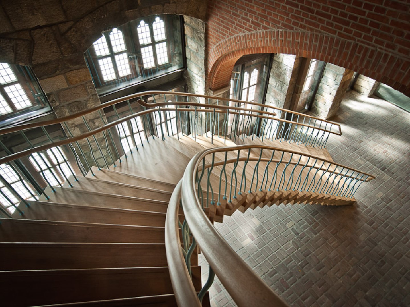 Grote houten trap met golvende blauwgrijze spijlen en houten leuning, in een monumentaal gebouw met bakstenen muren, natuurstenen elementen en hoge ramen.