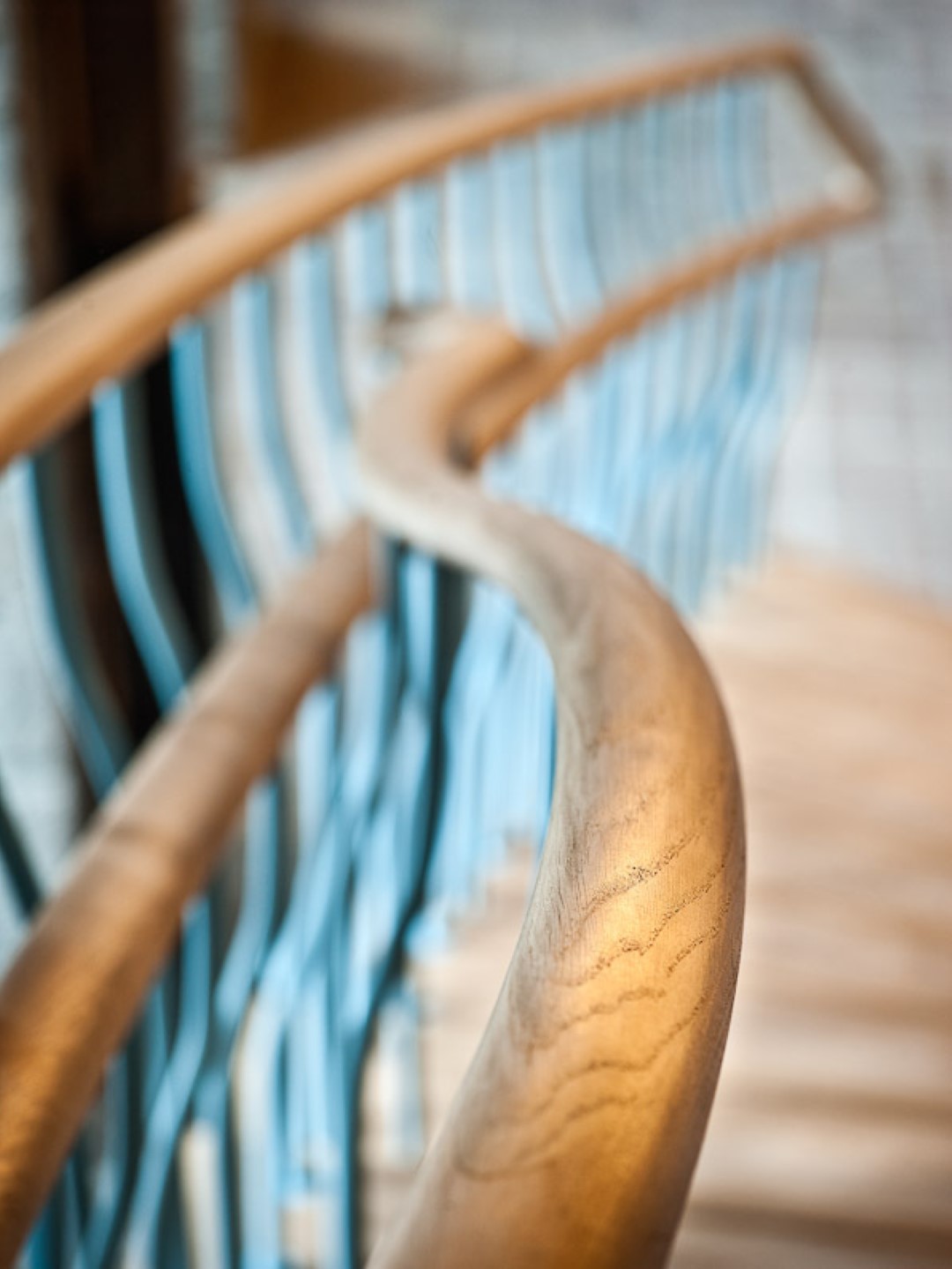 Close-up van een golvende houten trapleuning met daarachter blauwgrijze spijlen die een speels patroon vormen langs een houten trap.