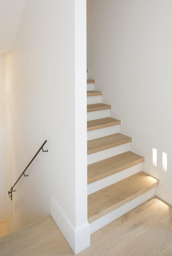 Een minimalistische trap met lichte houten treden en witte afwerking, geplaatst in een helder wit interieur; subtiele wandverlichting bij de onderste treden en een zwarte metalen leuning aan de trap naar beneden zorgen voor een moderne uitstraling.