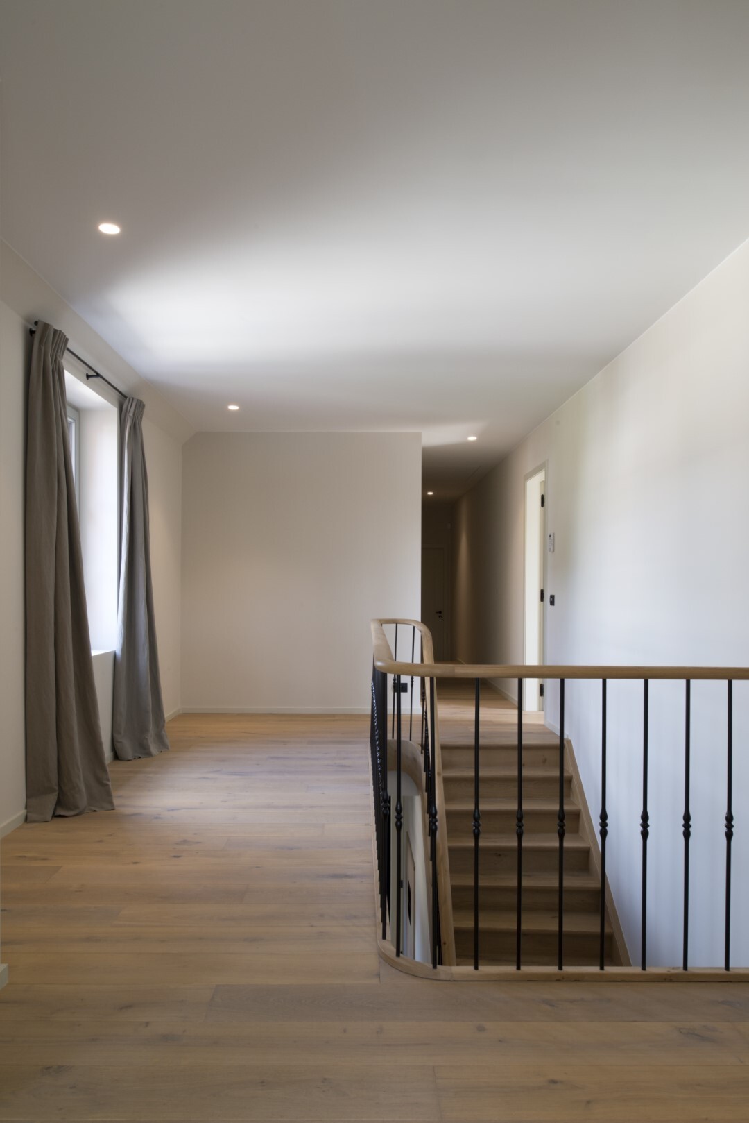 Eenvoudige overloop met lichte houten vloer, zwarte spijlenbalustrade rond de trapopening en een raam met grijze gordijnen links.
