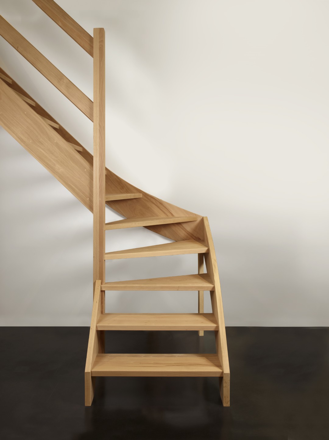 Een symmetrisch vooraanzicht van een lichte houten trap met open treden en een kwartdraai onderaan; de trap heeft een eenvoudige houten leuning en staat tegen een witte muur boven een donkere vloer.