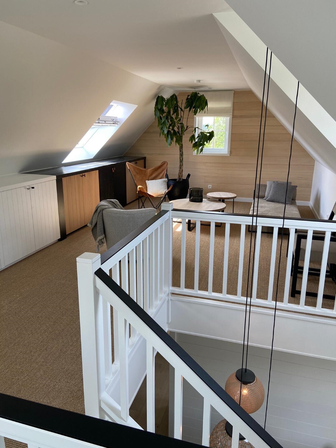 Gezellige zolderkamer met schuine plafonds, witte balustrade rond het trapgat, een zithoek met fauteuils en planten, en natuurlijke lichtinval via dakramen.