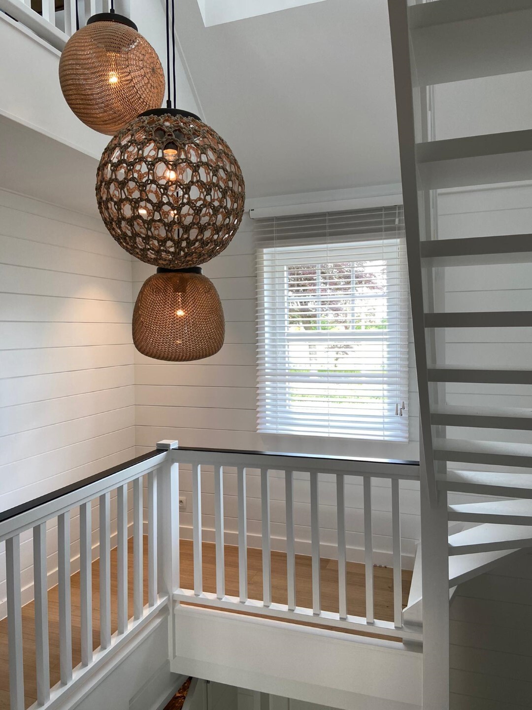 Lichte trappenhal met witte houten trap en balustrade, drie rotan hanglampen en een raam met jaloezieën dat natuurlijk licht binnenlaat.