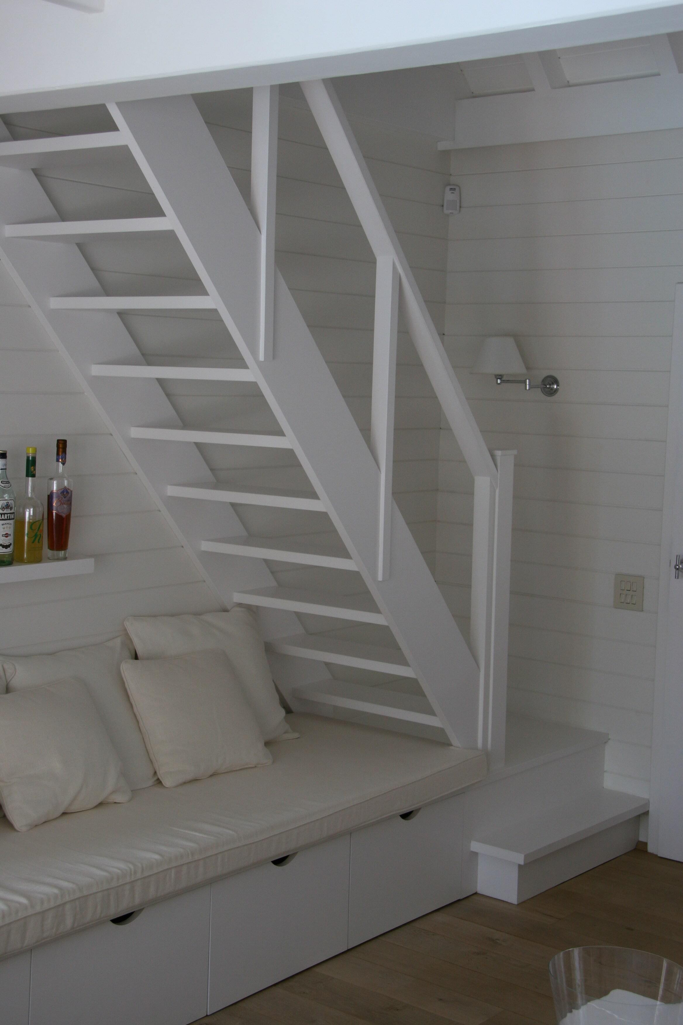 Een smalle witte open trap met verticale spijlen, geplaatst boven een ingebouwde zitbank met witte kussens, in een compacte, minimalistisch ingerichte woonruimte met witte houten wandbekleding.