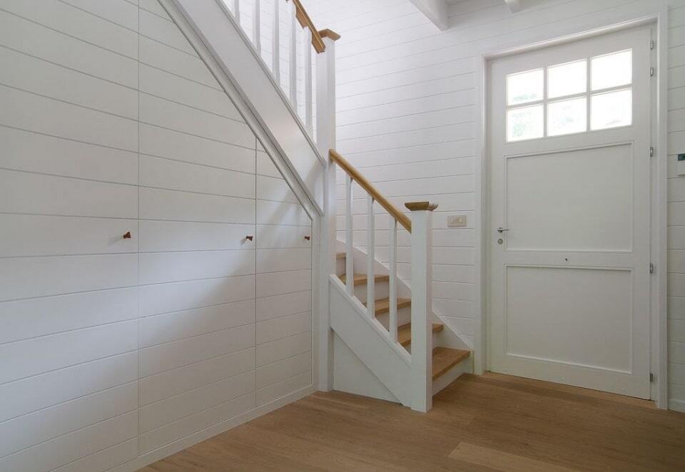 Een witgeschilderde houten trap met lichte houten treden en bijpassende leuning staat in een landelijke hal met witgelambriseerde wanden, een houten vloer en een witte paneeldeur met glasroeden.
