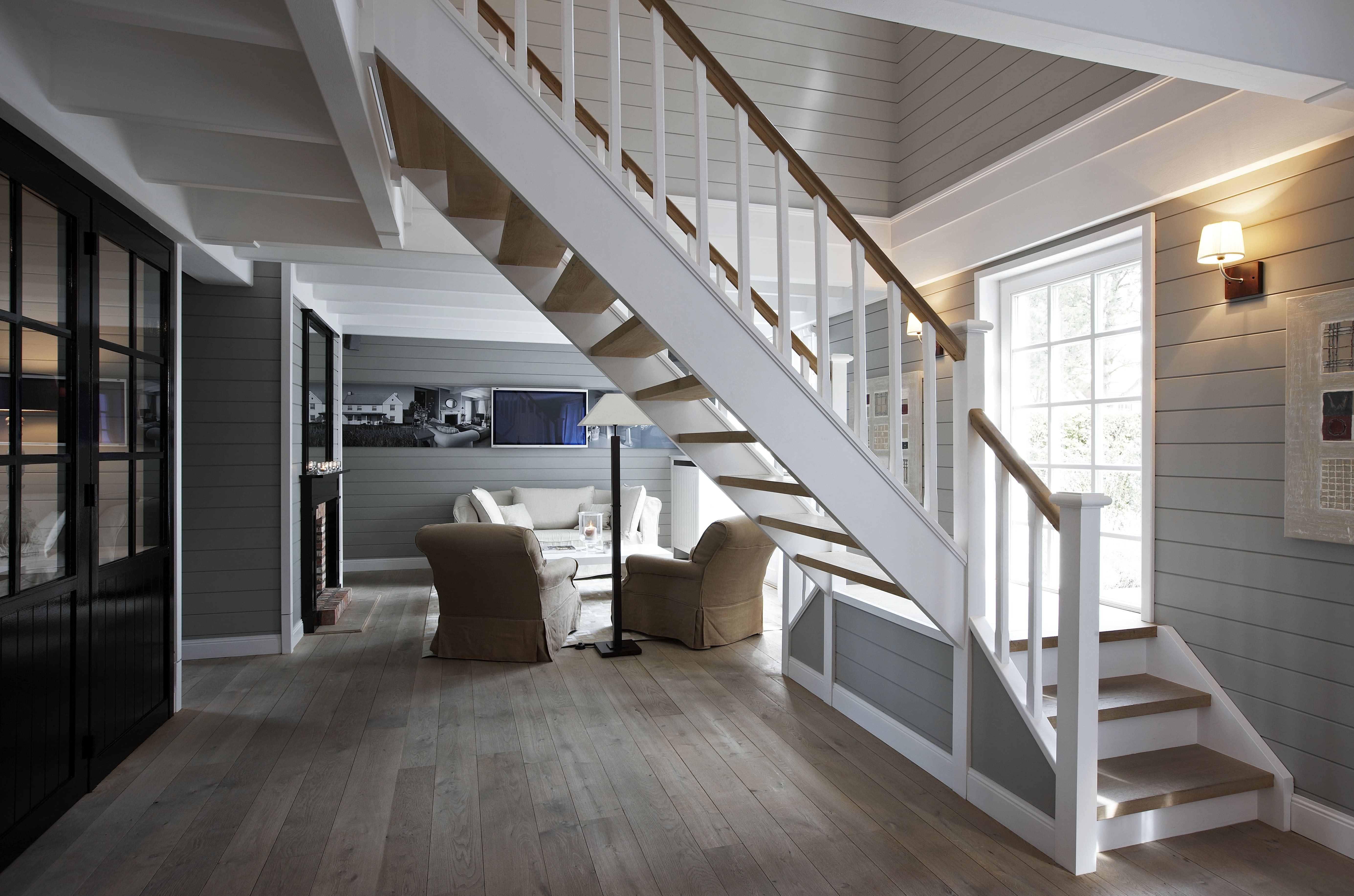 Een witgelakte houten trap met naturel houten treden en spijlenleuning staat centraal in een ruime, sfeervolle woonruimte met grijs lambrisering, grote ramen, een houten vloer en een gezellige zithoek met beige fauteuils op de achtergrond.