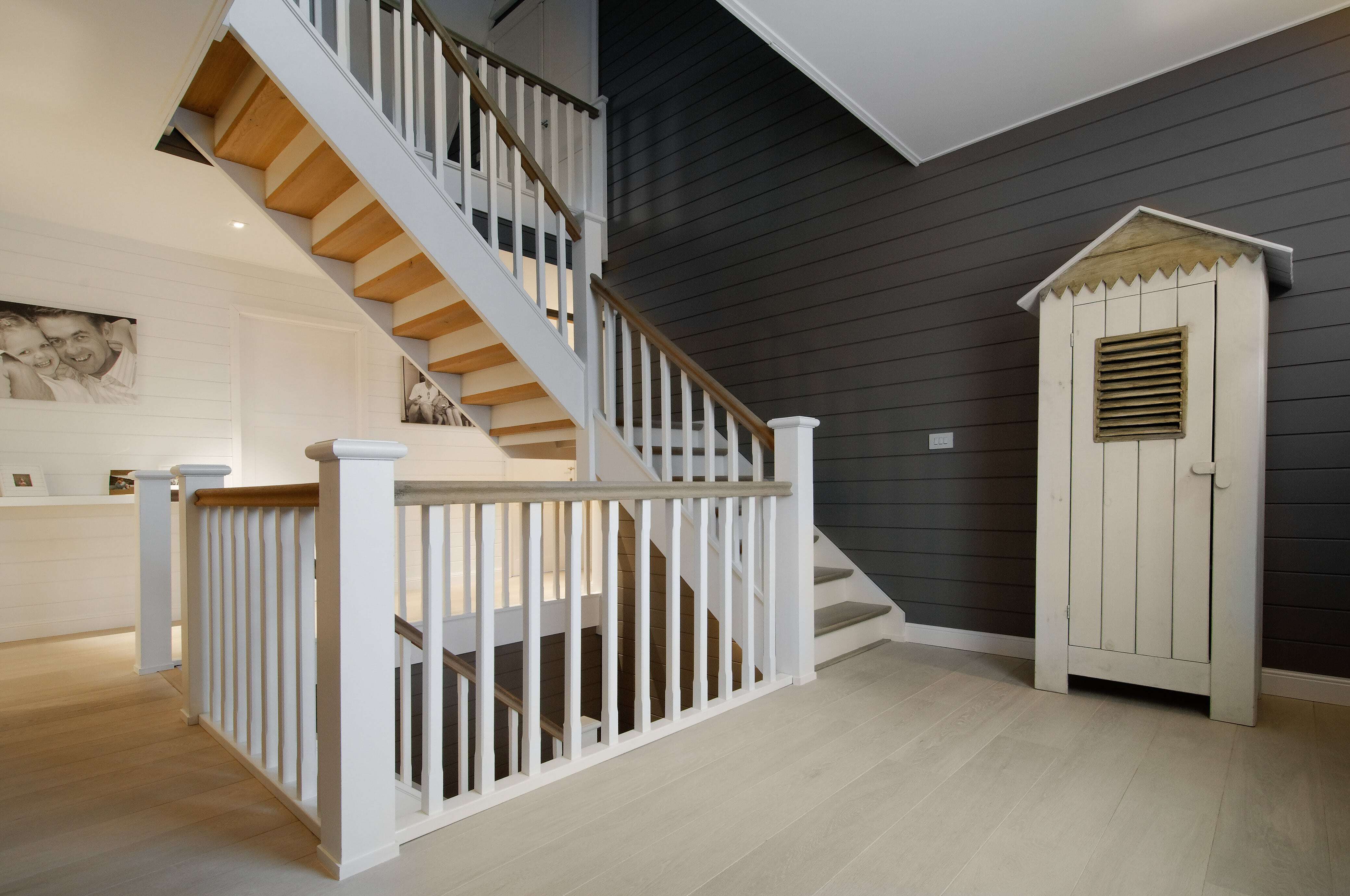 Een robuuste witte houten trap met naturel treden, spijlenbalustrade en een centraal bordes staat in een modern-landelijke woning met licht houten vloer, donkergrijze lambrisering en een speels kastje in de vorm van een strandhuisje.