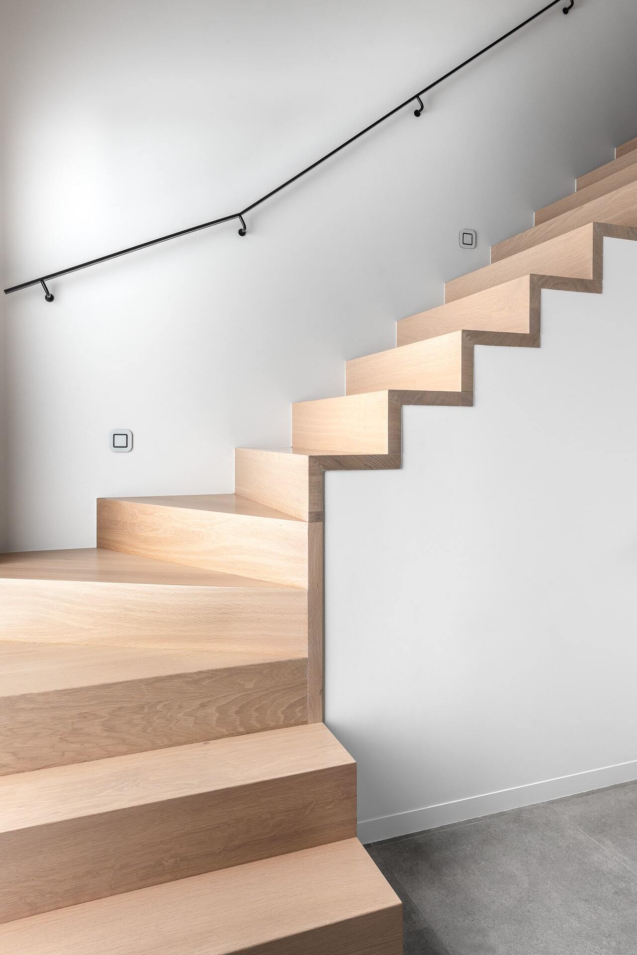 Een moderne houten trap met een opvallend zigzagprofiel, strak gemonteerd tegen een witte muur, zonder leuning, met een minimalistische en zwevende uitstraling.