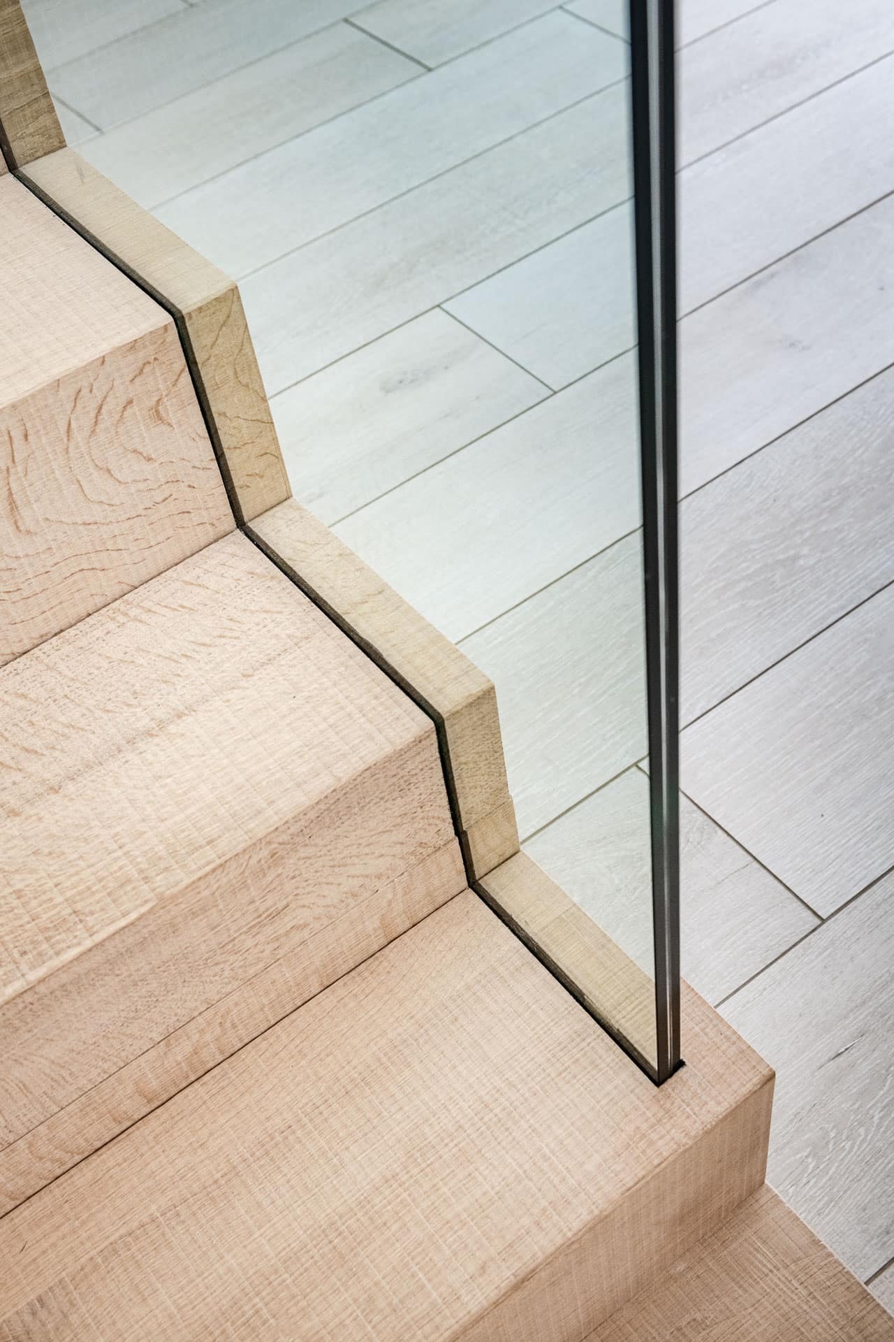 Close-up van lichte houten traptreden met een strakke zwarte belijning en een glazen balustrade, overgaand in een vloer met brede, lichte planken.