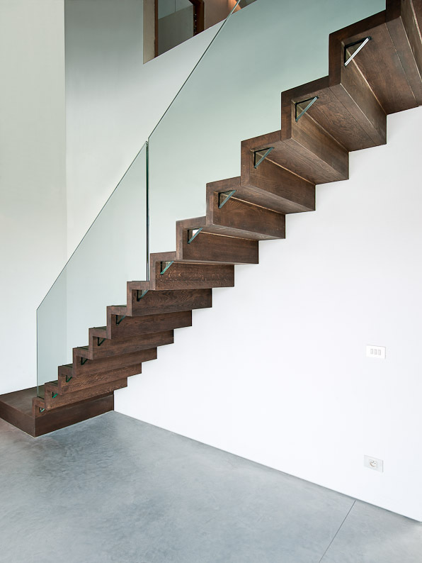 Moderne trap met donkerhouten zigzagtreden en een doorlopende glazen balustrade, geplaatst langs een witte muur boven een grijze gietvloer.