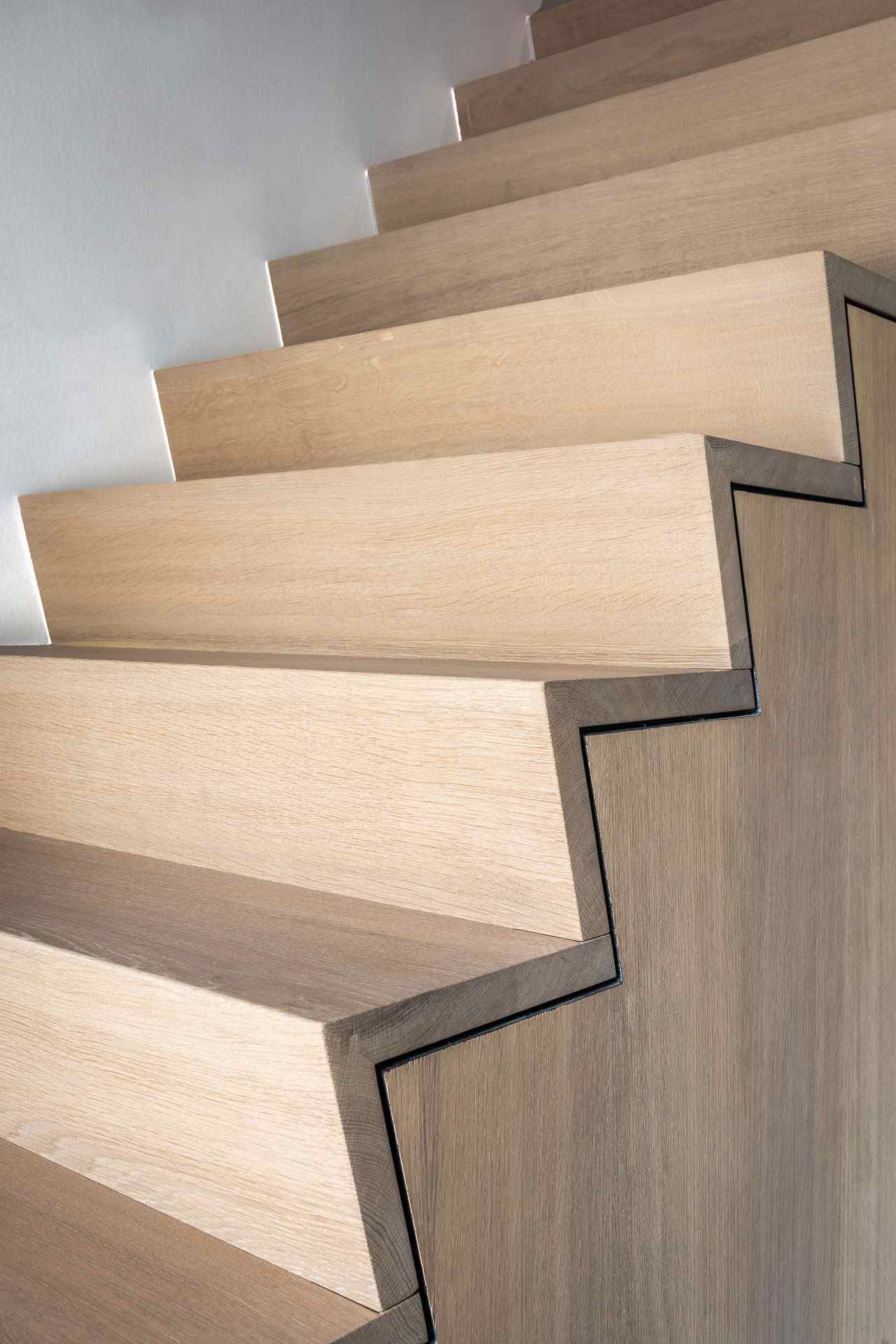 Detail van een moderne houten trap met scherpe zigzagvorm en zwarte lijnen die de treden visueel benadrukken tegen een lichte achtergrond.