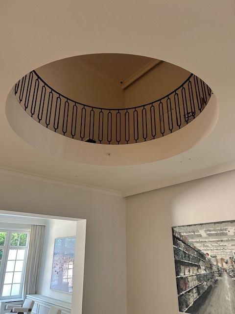 Een cirkelvormige vide in het plafond met een zwarte sierlijke balustrade, in een lichte kamer met kunst aan de muur en een raam met zicht op buiten.
