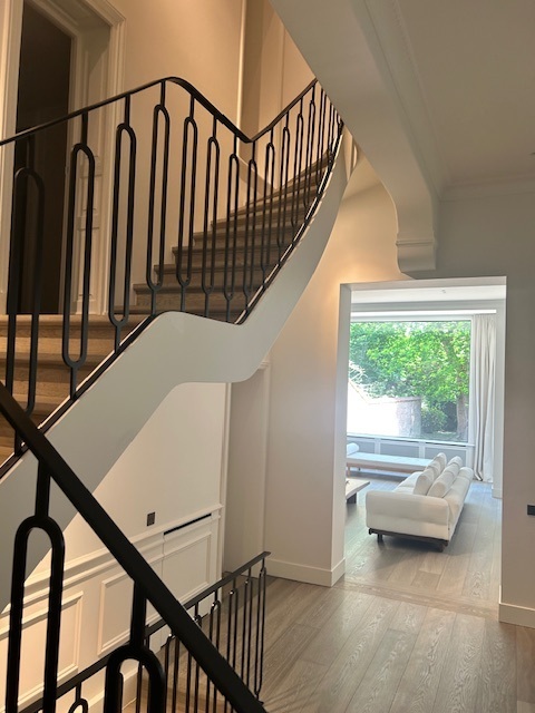 Een elegante trap met zwarte sierleuning en witte zijkant, met zicht op een lichte woonkamer en groene tuin door een open deuropening.