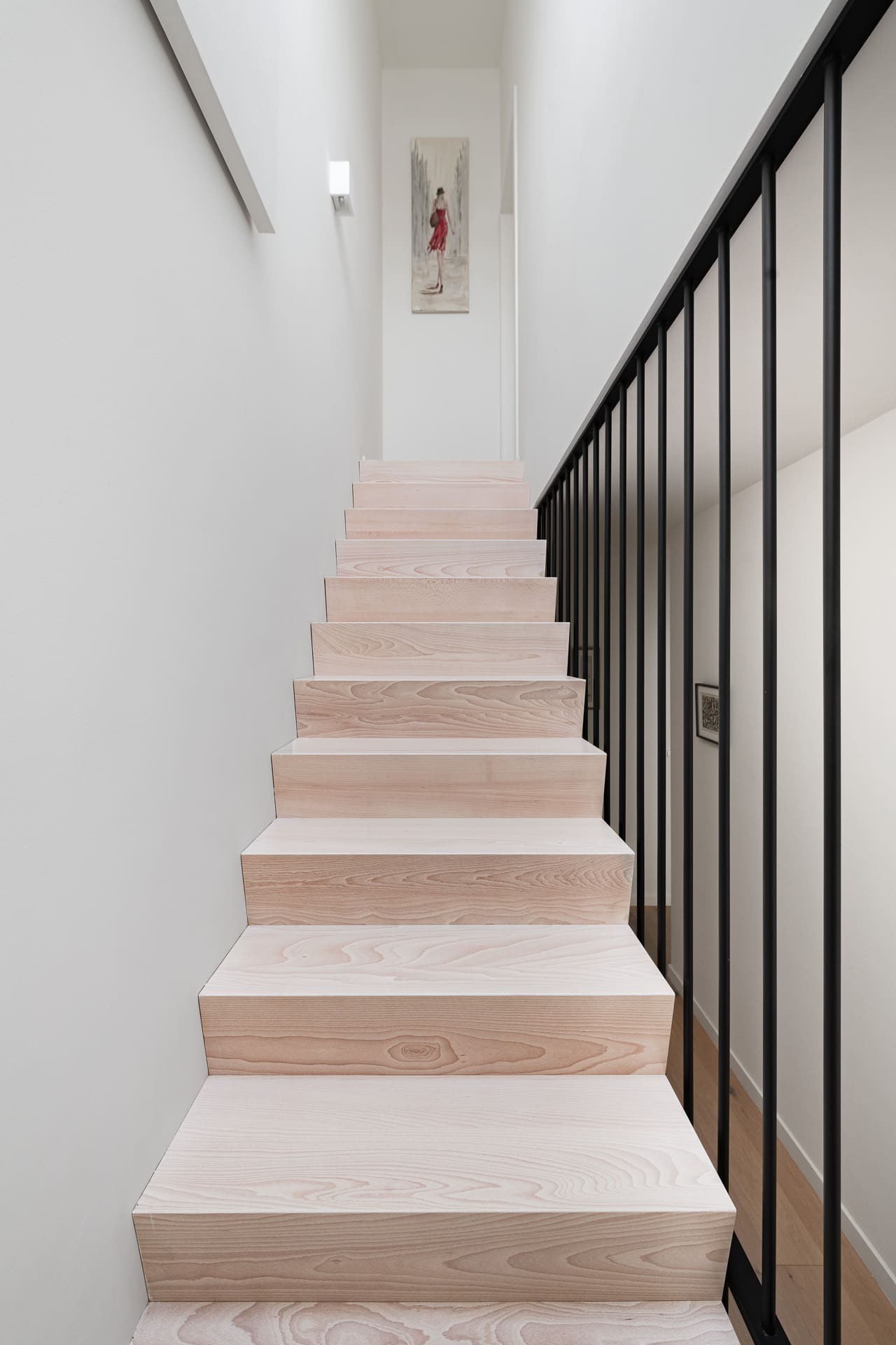 Rechte trap met gesloten lichte houten treden, witte muren, een zwarte verticale balustrade en een kunstwerk aan het einde van de gang.