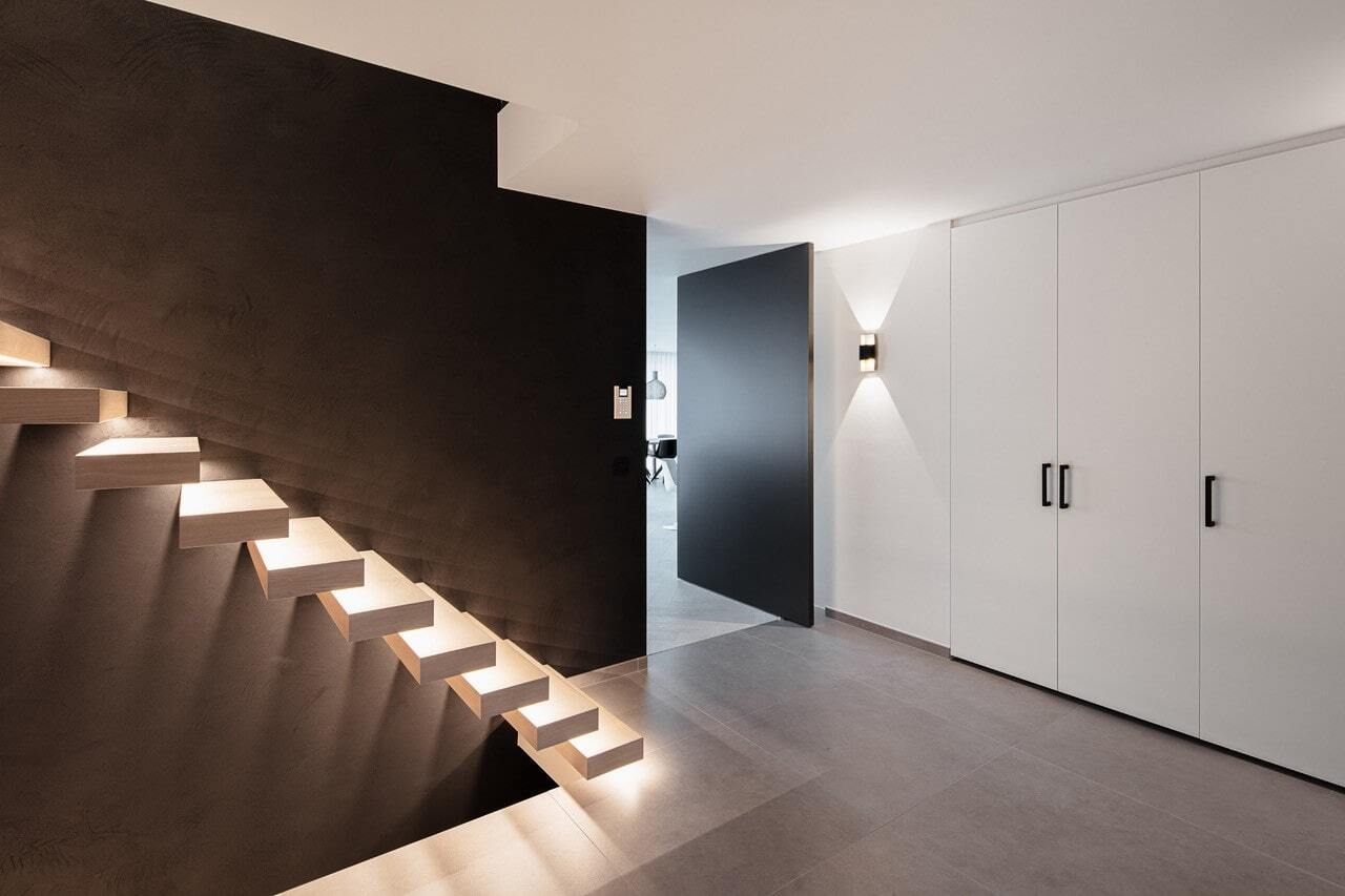 Strak vormgegeven hal met een zwarte muur, zwevende houten trap met geïntegreerde ledverlichting, witte kastenwand en een open zwarte deur richting een lichtere ruimte.