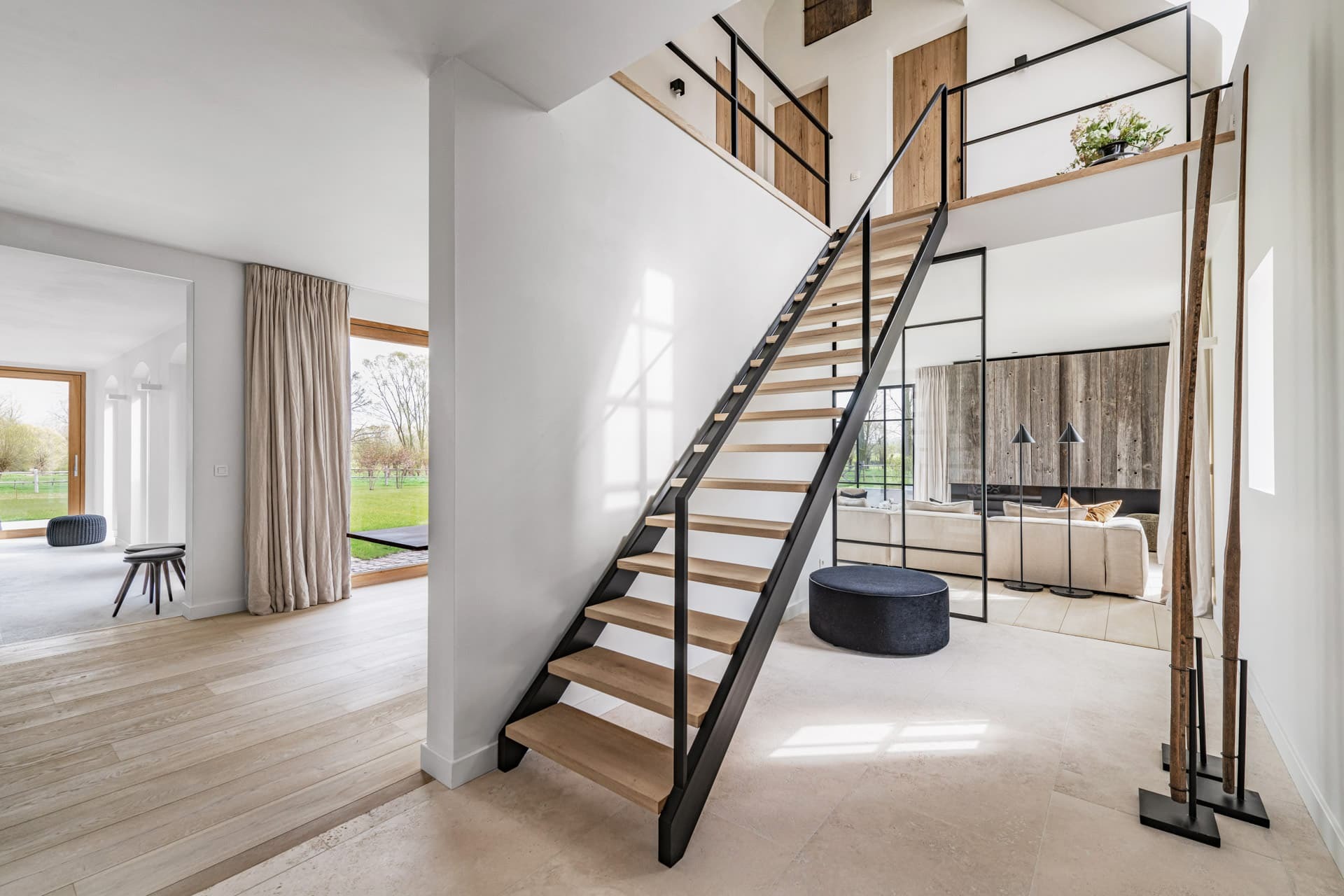 Ruime en lichte woonkamer met een open houten trap met zwarte leuningen, grote ramen, een zwarte poef, lichte vloeren en een houten deur op de bovenverdieping.
