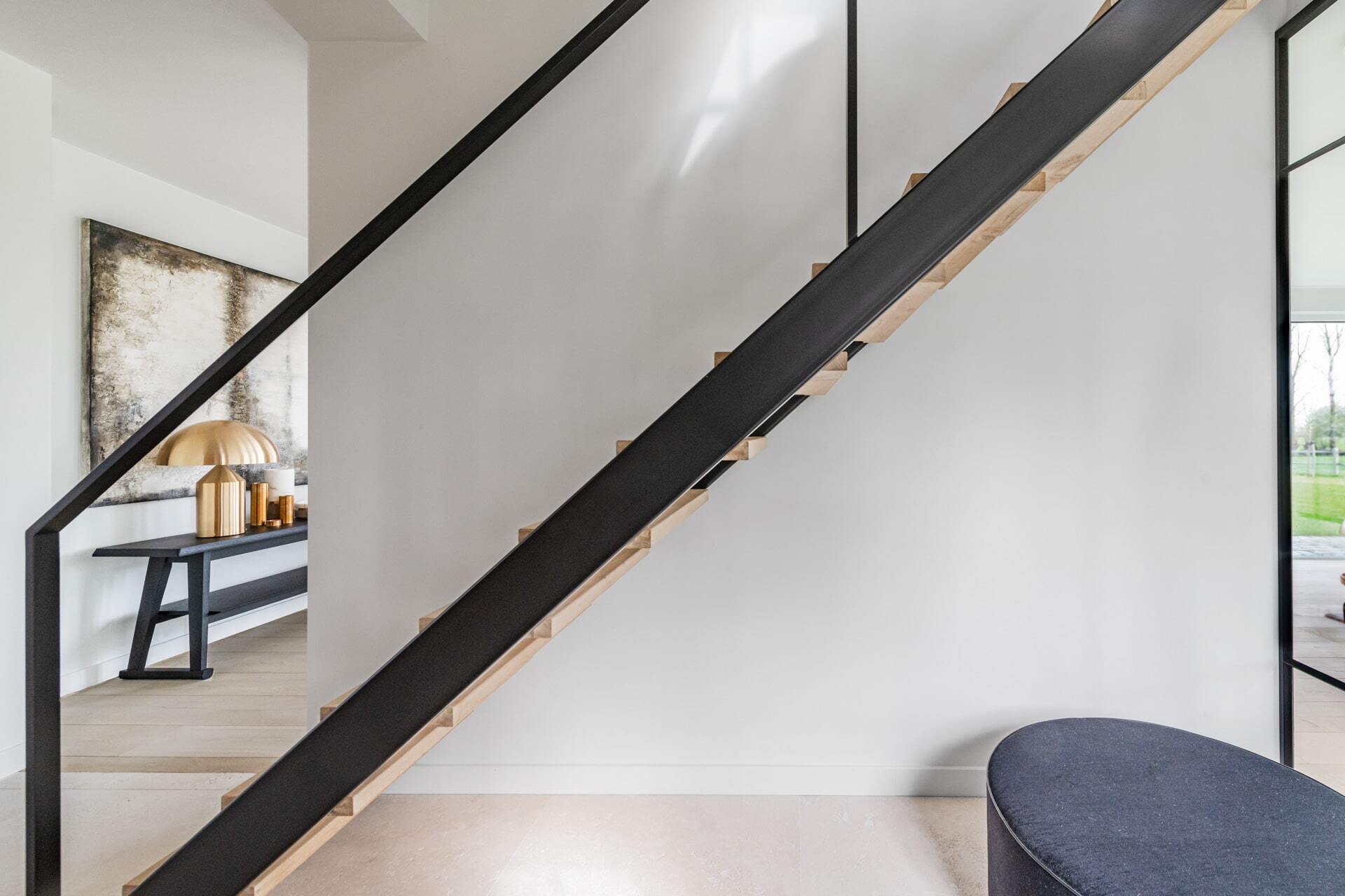 Zwarte trap met houten treden in een modern, minimalistisch interieur met witte muren en een donkere poef op de voorgrond.