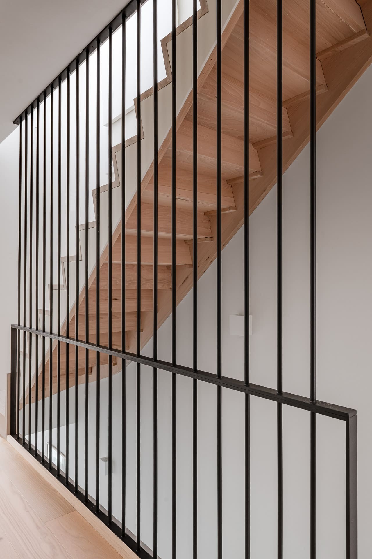 Zijaanzicht van de onderzijde van een trap met gesloten houten treden, omringd door een hoge zwarte spijlenbalustrade in een strak, modern interieur.