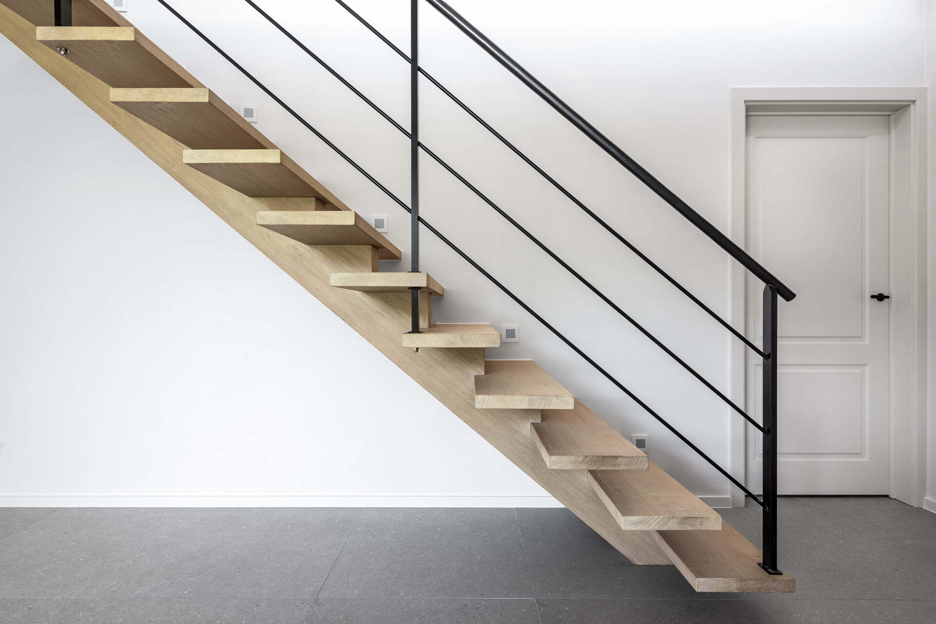 Zijaanzicht van een moderne open trap met lichte houten treden en zwarte metalen balustrade, geplaatst tegen een witte muur met een gesloten witte deur ernaast.