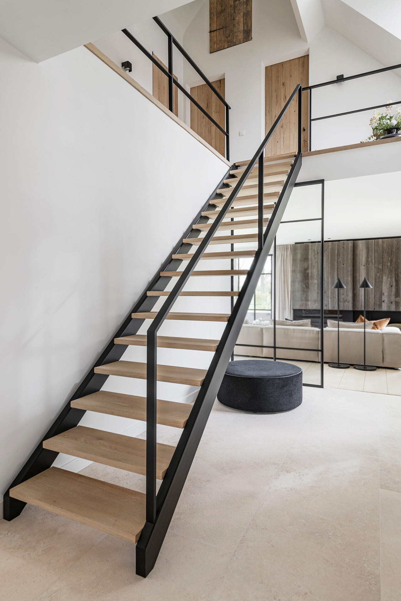 Rechte open trap met houten treden en zwarte leuningen in een licht en modern interieur, met een zwarte poef en houten deuren op de bovenverdieping.
