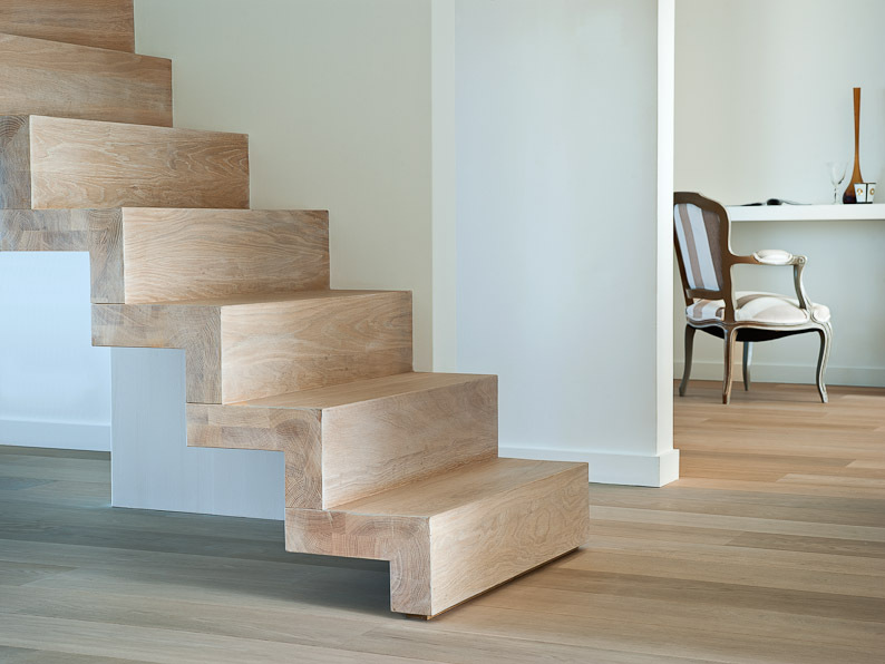 Lichte houten trap met gesloten treden, geplaatst in een warm interieur met houten vloer en een klassieke stoel zichtbaar in de naastgelegen ruimte.