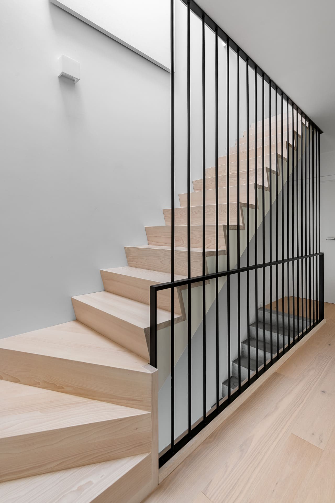 Moderne trap met gesloten lichte houten treden en een opvallende zwarte spijlenbalustrade die tot aan het plafond reikt, in een strak wit interieur.