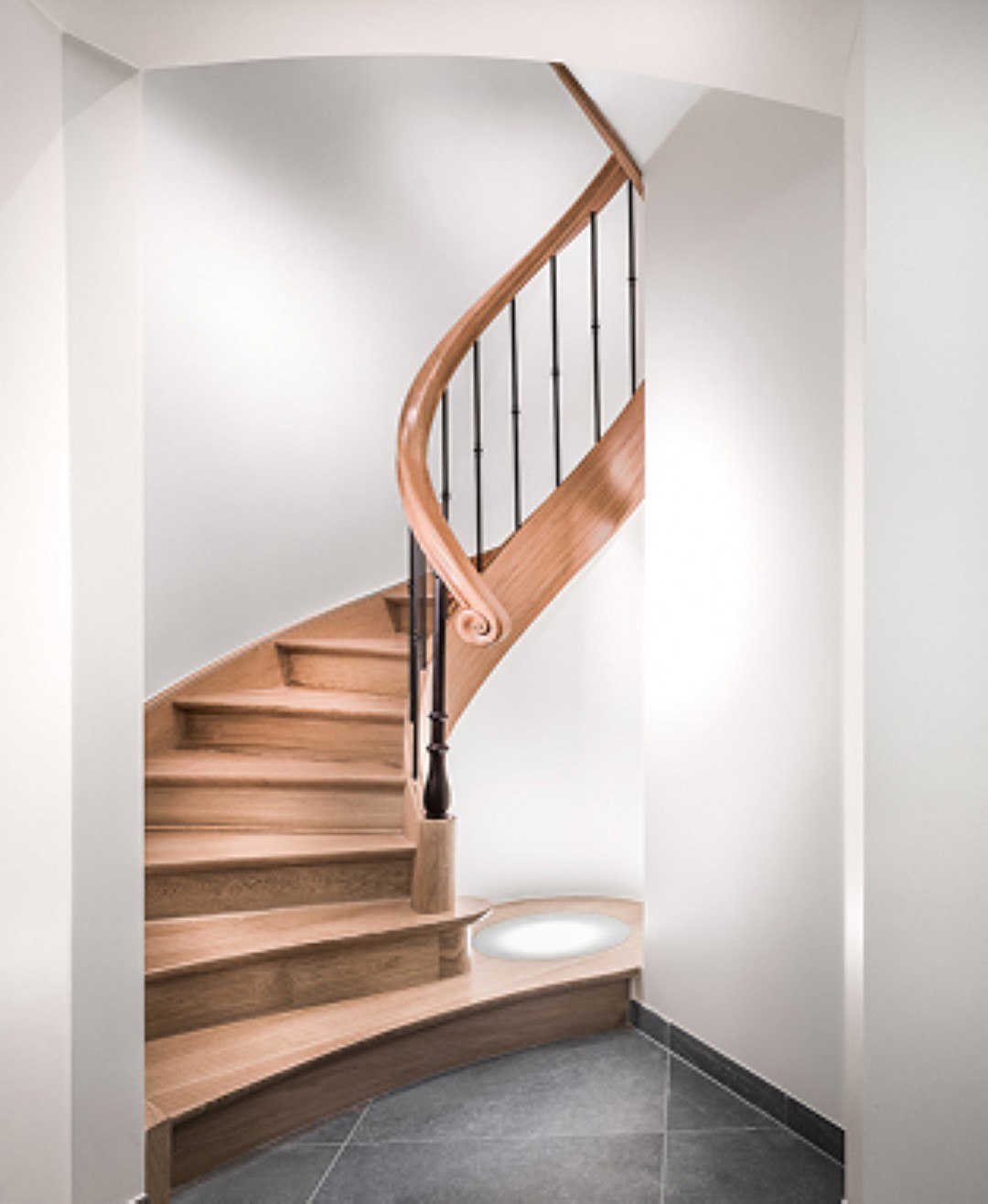 Elegante houten draaitrap met sierlijke leuning en zwarte spijlen, geplaatst in een smalle nis met witte muren en grijze tegelvloer.