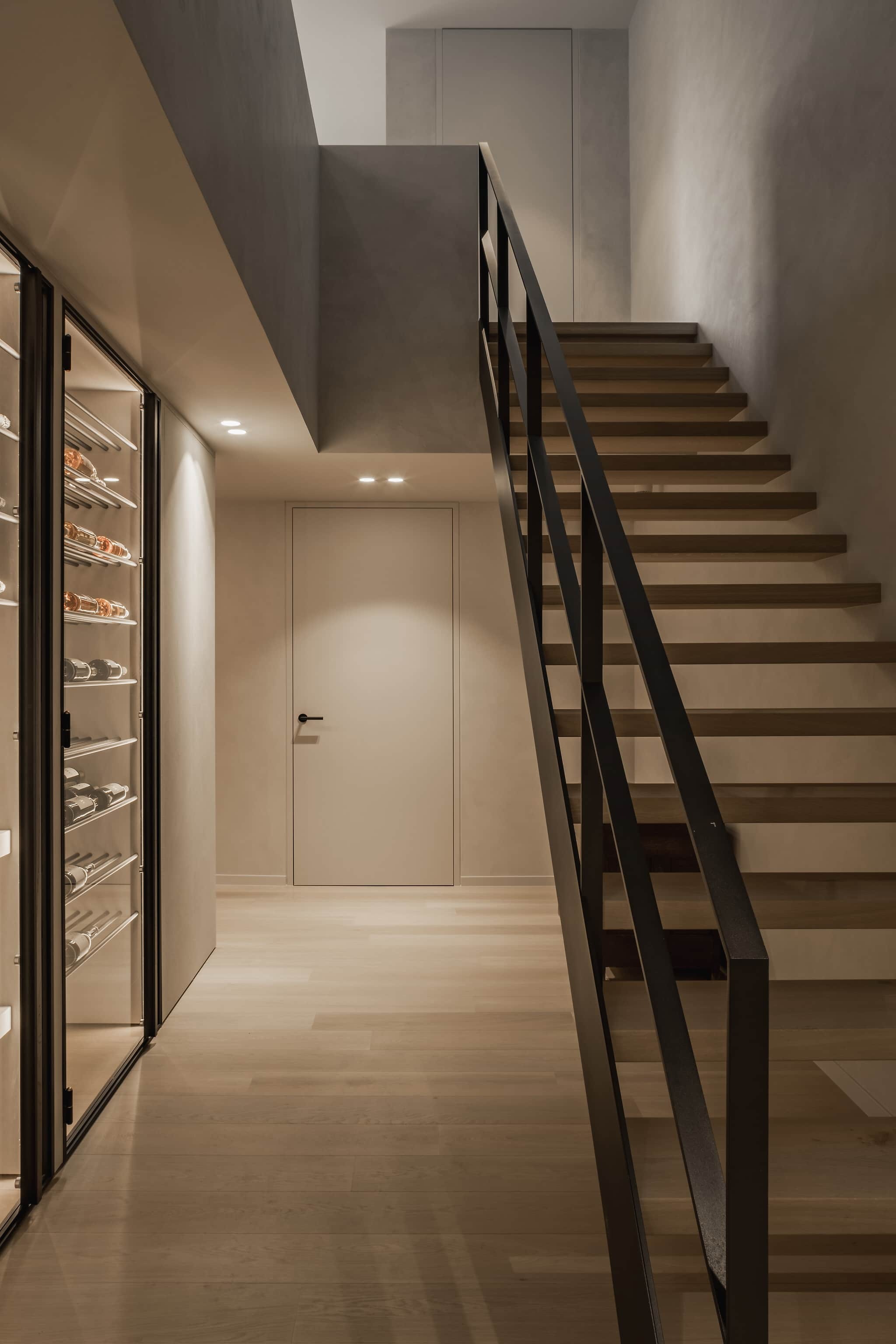 Stijlvolle ruimte met een zwevende houten trap met zwarte leuning, een glazen wijnkast langs de wand, warme verlichting en een houten vloer die doorloopt naar een gesloten deur.